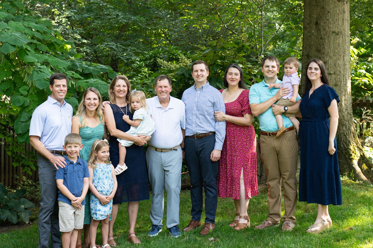 Large family group portrait outdoors, smiling adults and children posing together on green lawn with trees.