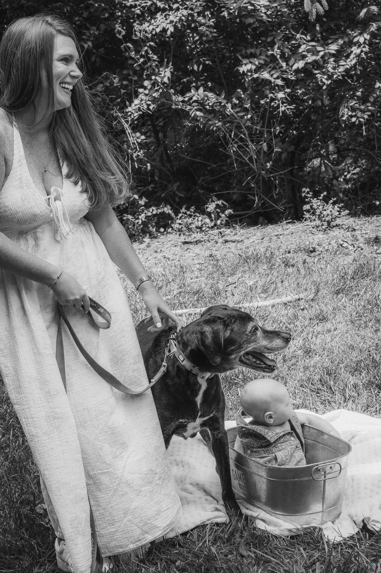 Black and white photo of smiling woman in white dress holding dog leash outdoors with baby in metal bucket.