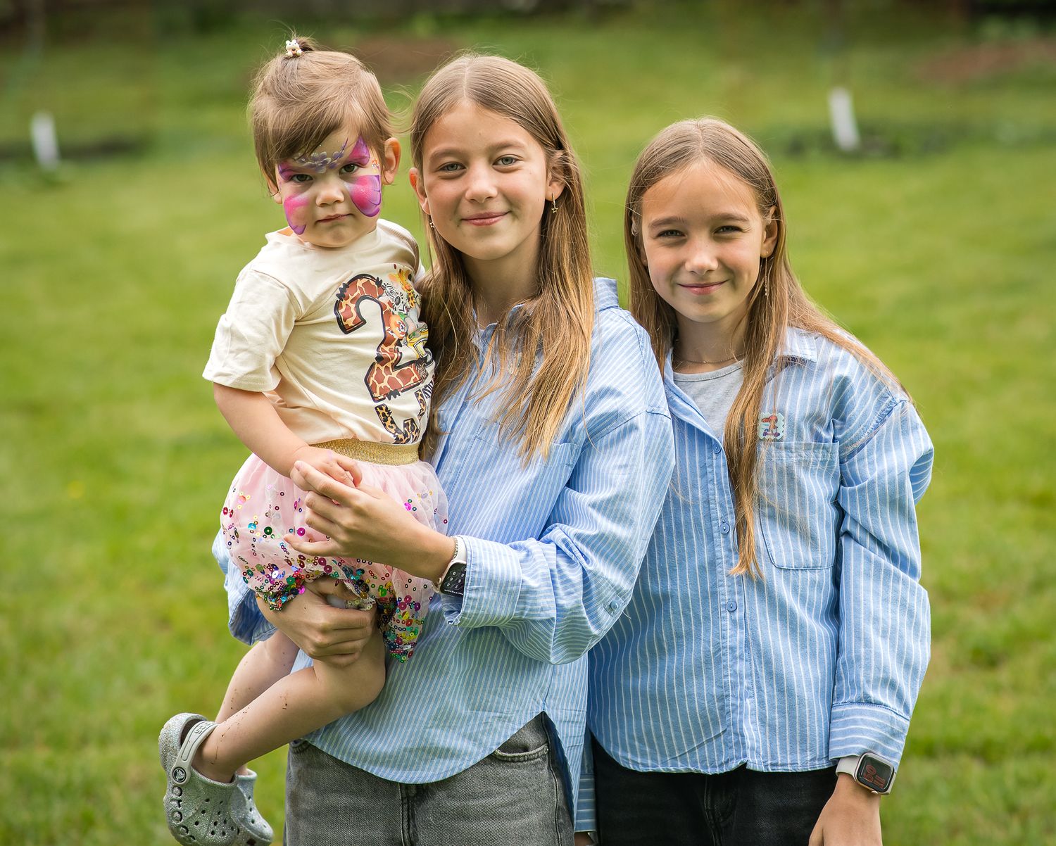 Three young girls smiling outdoors, oldest holding toddler with face paint, standing on green grass.