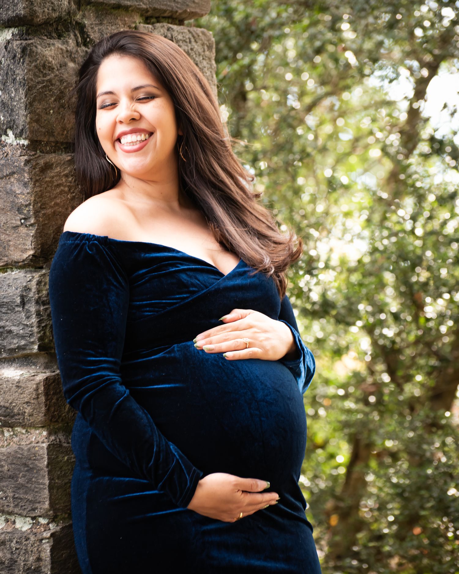 Smiling pregnant woman in navy blue velvet off-shoulder dress cradling baby bump outdoors near stone wall.