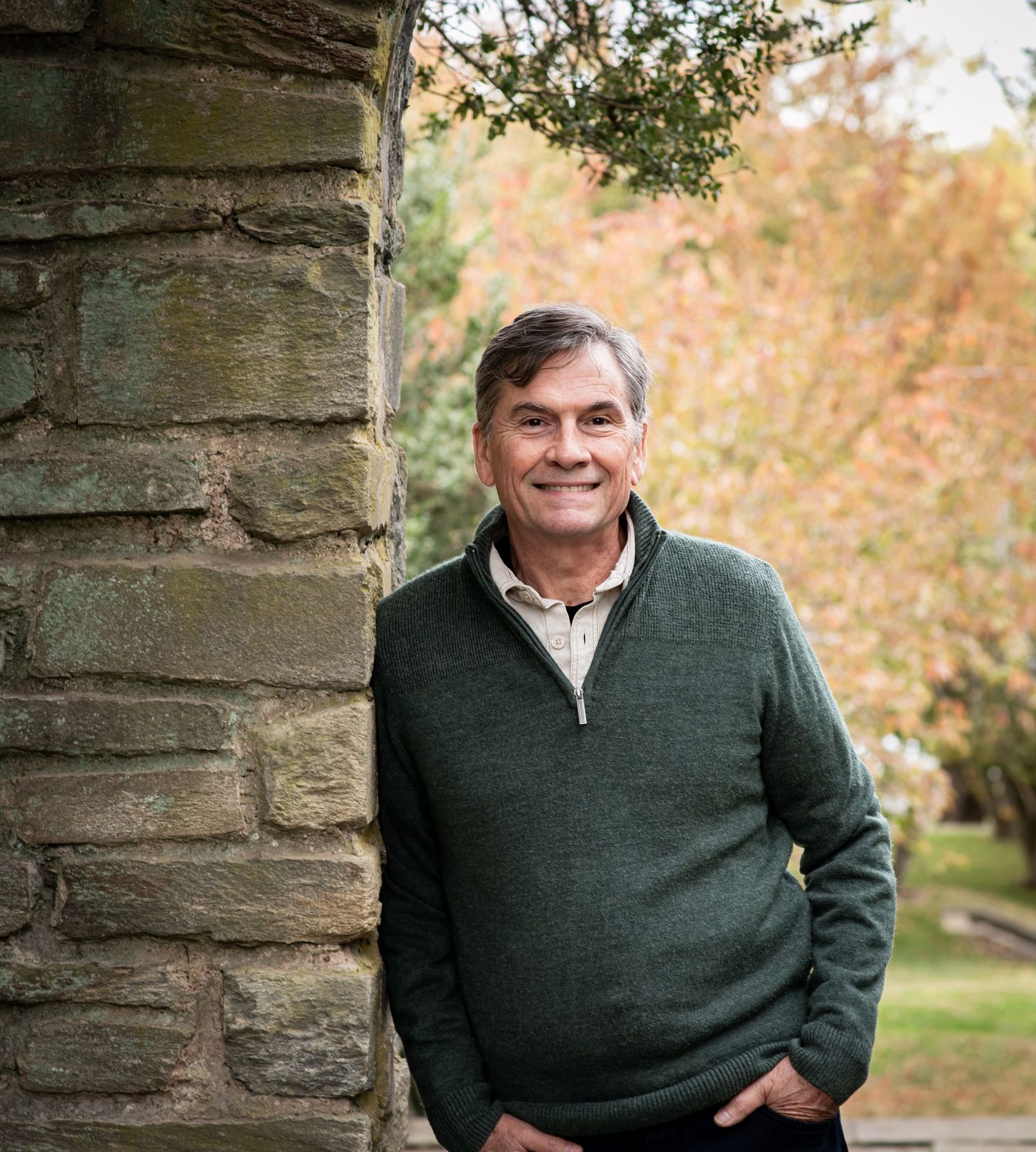 Person in a dark green sweater smiles warmly while posing against stone pillars in an autumn outdoor setting.