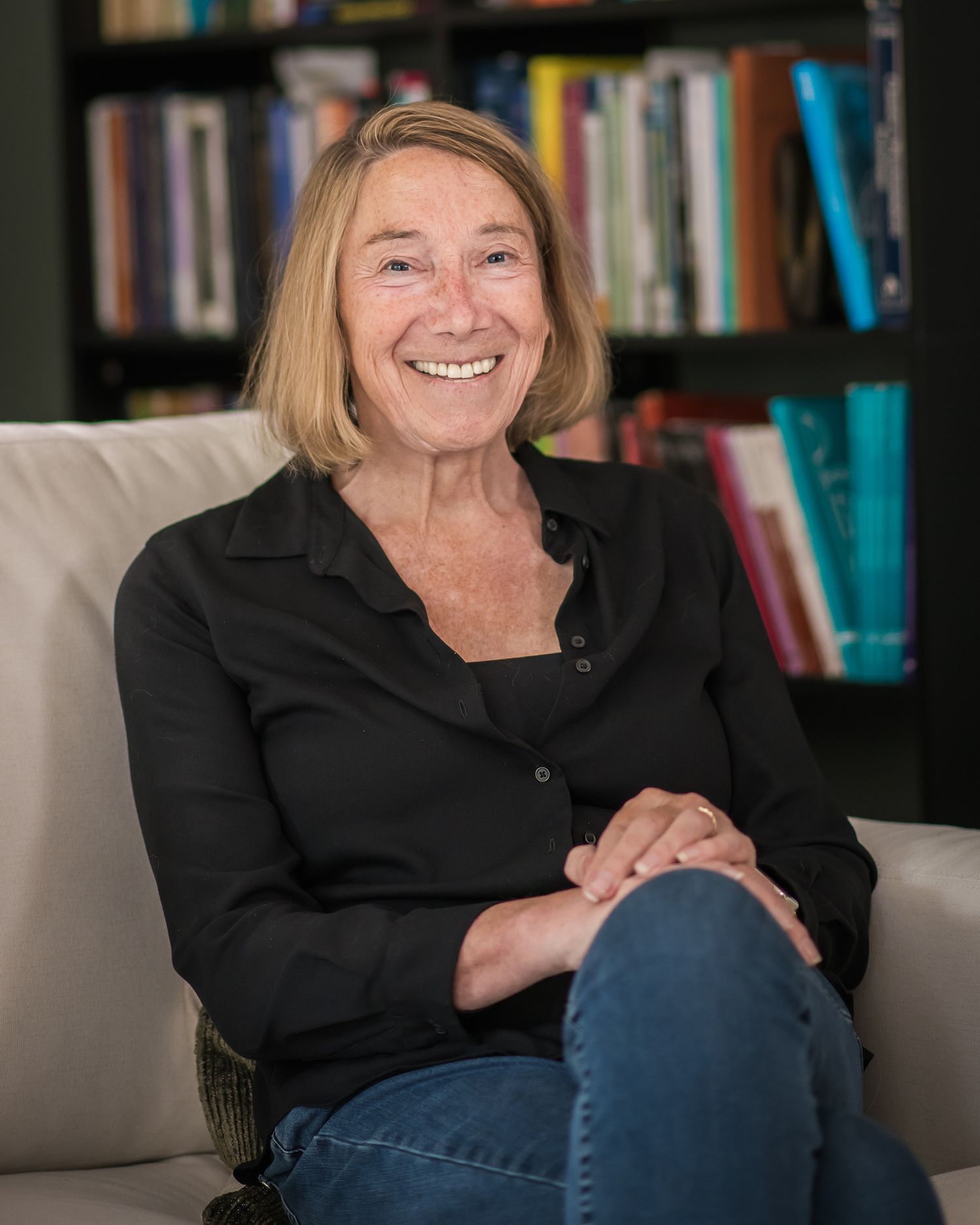 Smiling middle-aged woman with blonde hair sitting on a couch in front of a bookshelf, wearing a black shirt and jeans.