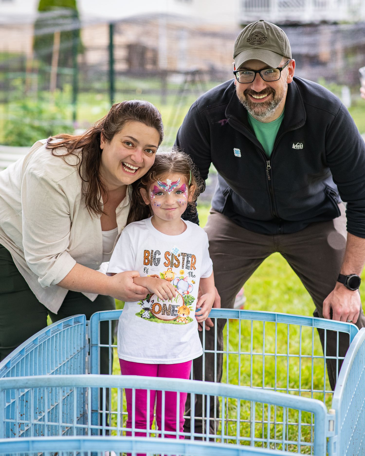 Family smiling with face-painted girl wearing Big Sister shirt at outdoor animal pen.