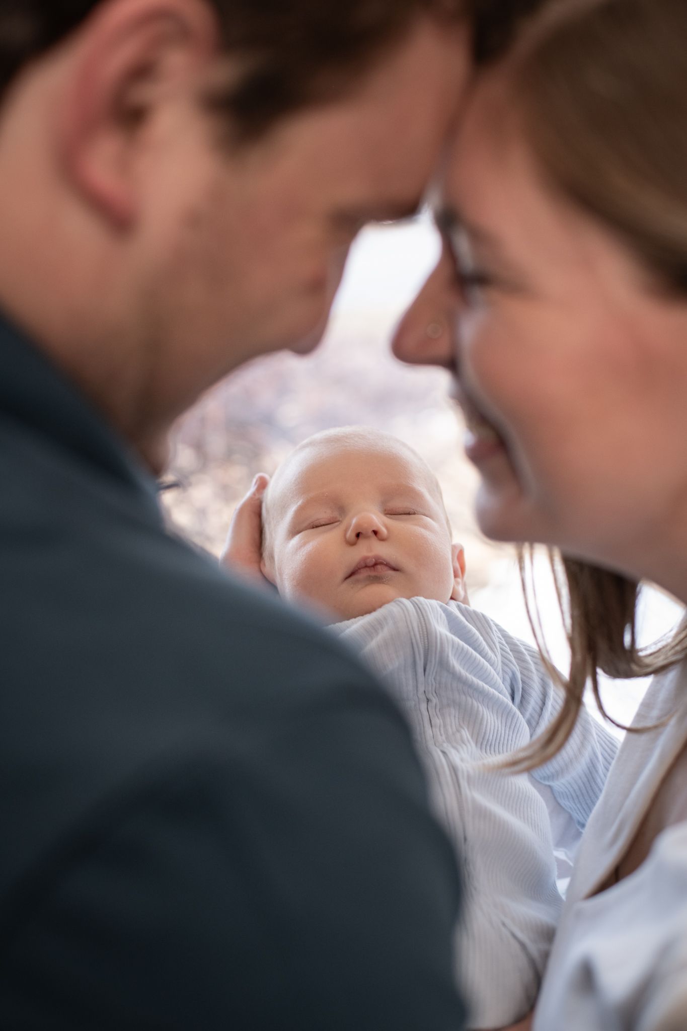 Parents gaze lovingly at their sleeping newborn baby wearing a white onesie in a tender family moment.