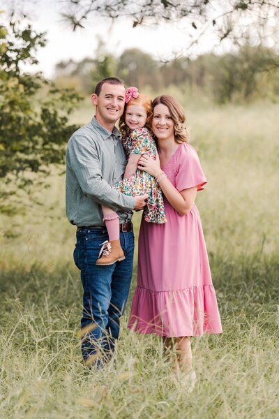 A family of three poses together in a sunny meadow wearing coordinated pink and blue outfits for outdoor portraits.