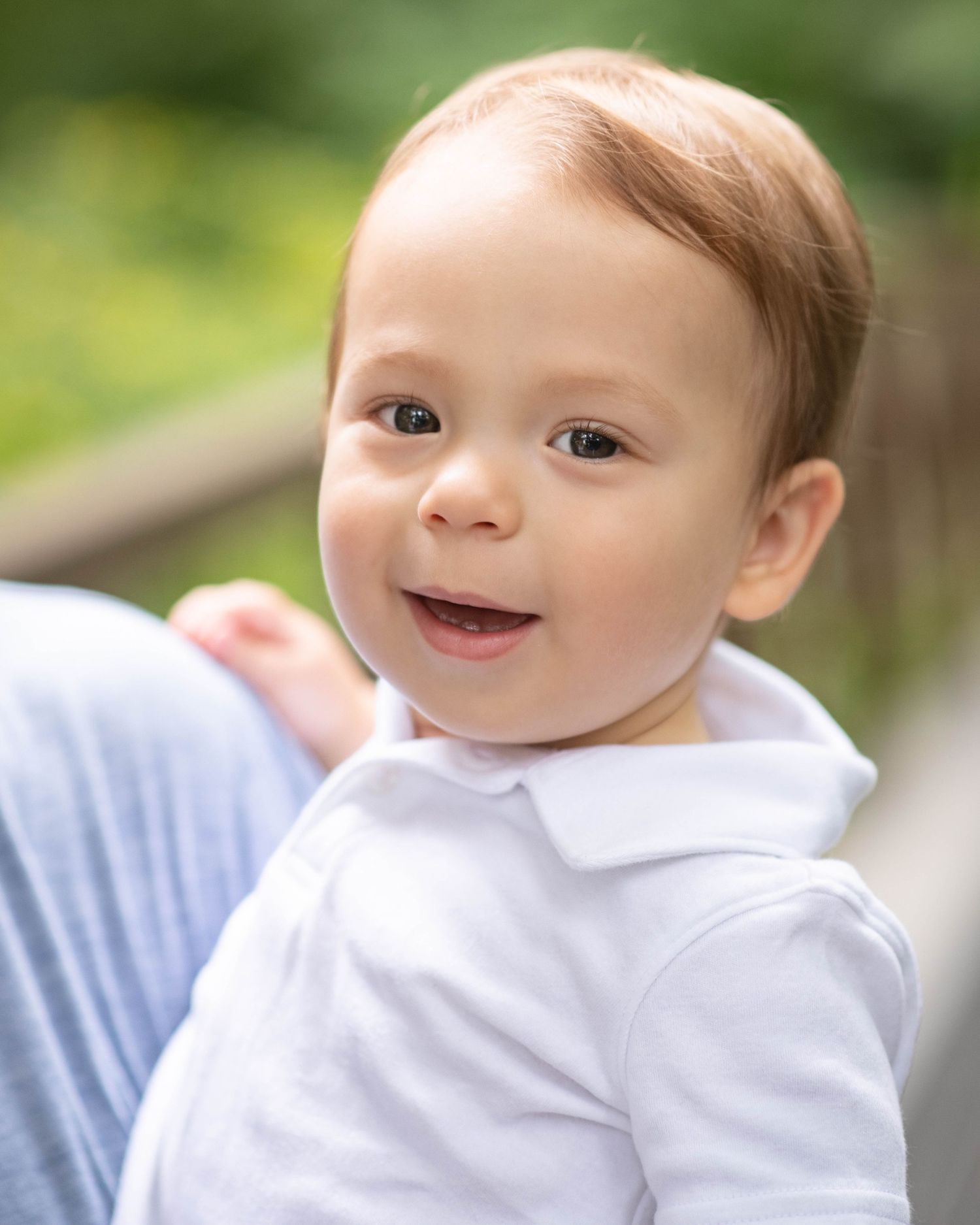 Smiling baby boy in white polo shirt with light brown hair, photographed outdoors in a bright garden setting.