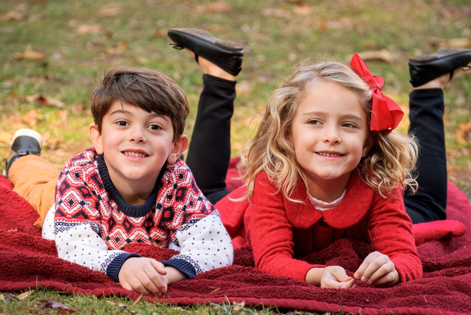 Two children lying on red blanket outdoors during autumn photo session.