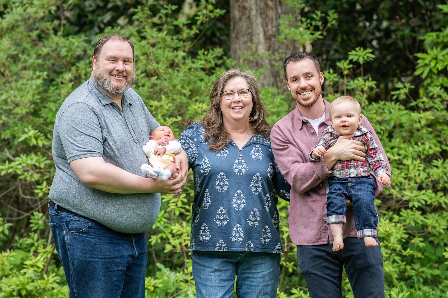 Family portrait with two men holding babies smiling outdoors in front of lush green trees.