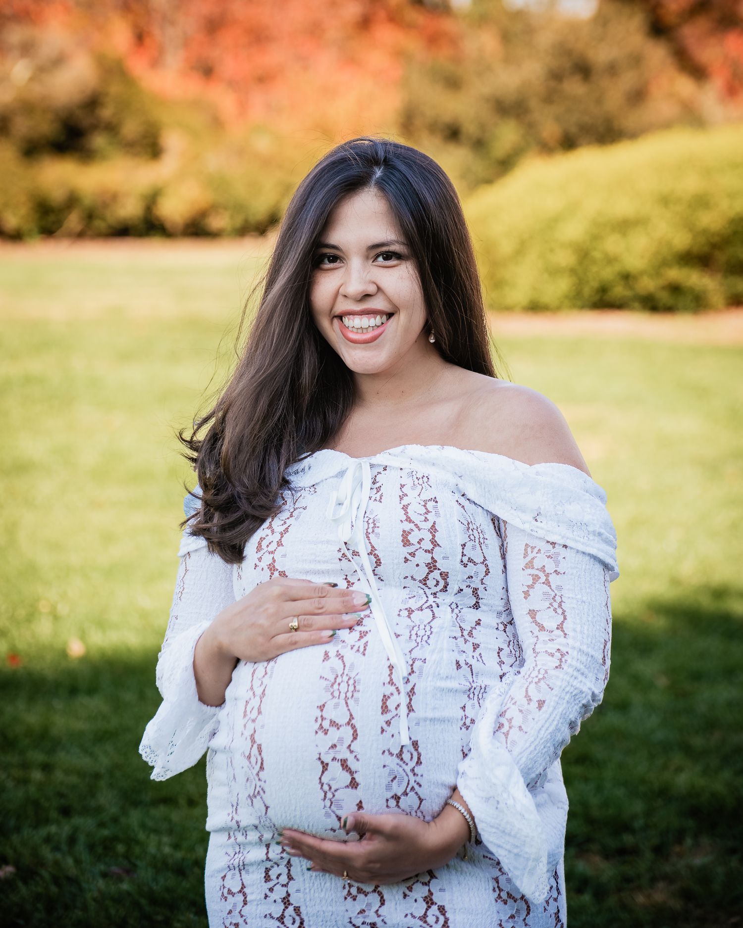Smiling pregnant woman in white lace off-shoulder dress cradling baby bump outdoors with autumn foliage background.