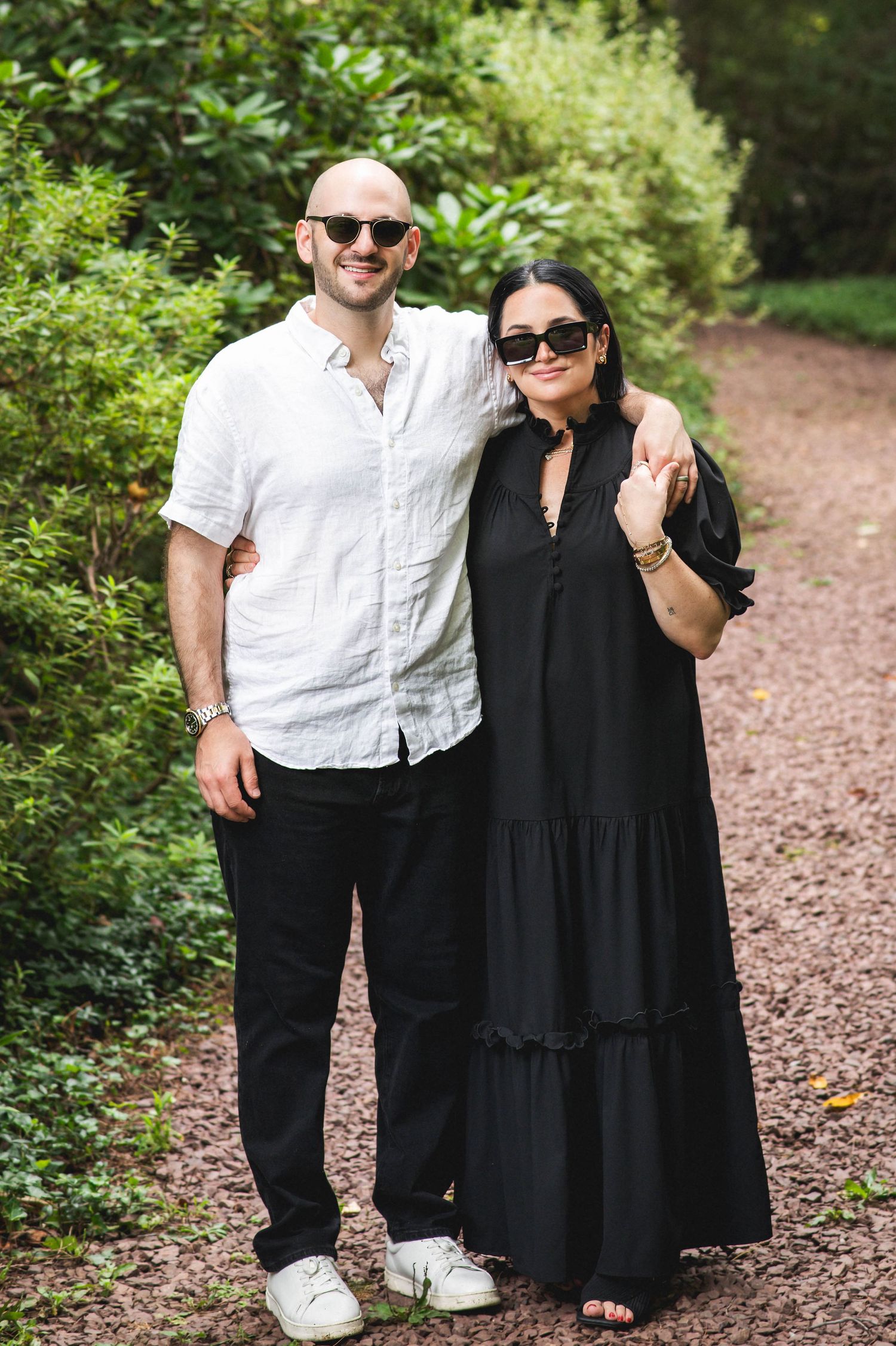 A bald man in white shirt and a woman in black dress pose together on a garden path wearing sunglasses.