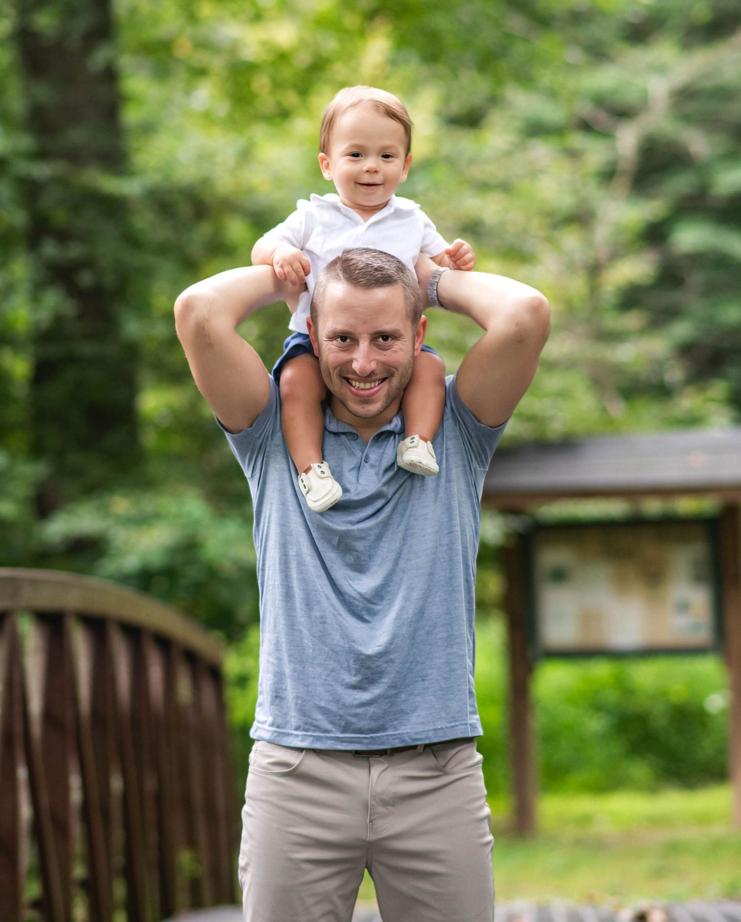 A smiling man carries a happy toddler on his shoulders outdoors in a lush green park setting.