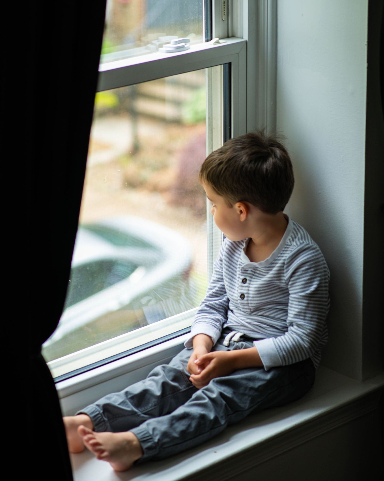 A young barefoot boy in striped pajamas sits on a window sill gazing outside on a rainy day.