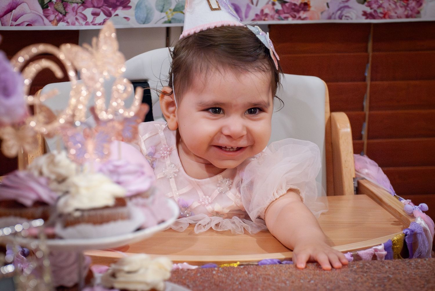 A child in a pink dress sits at a wooden table with a birthday cake and party decorations.