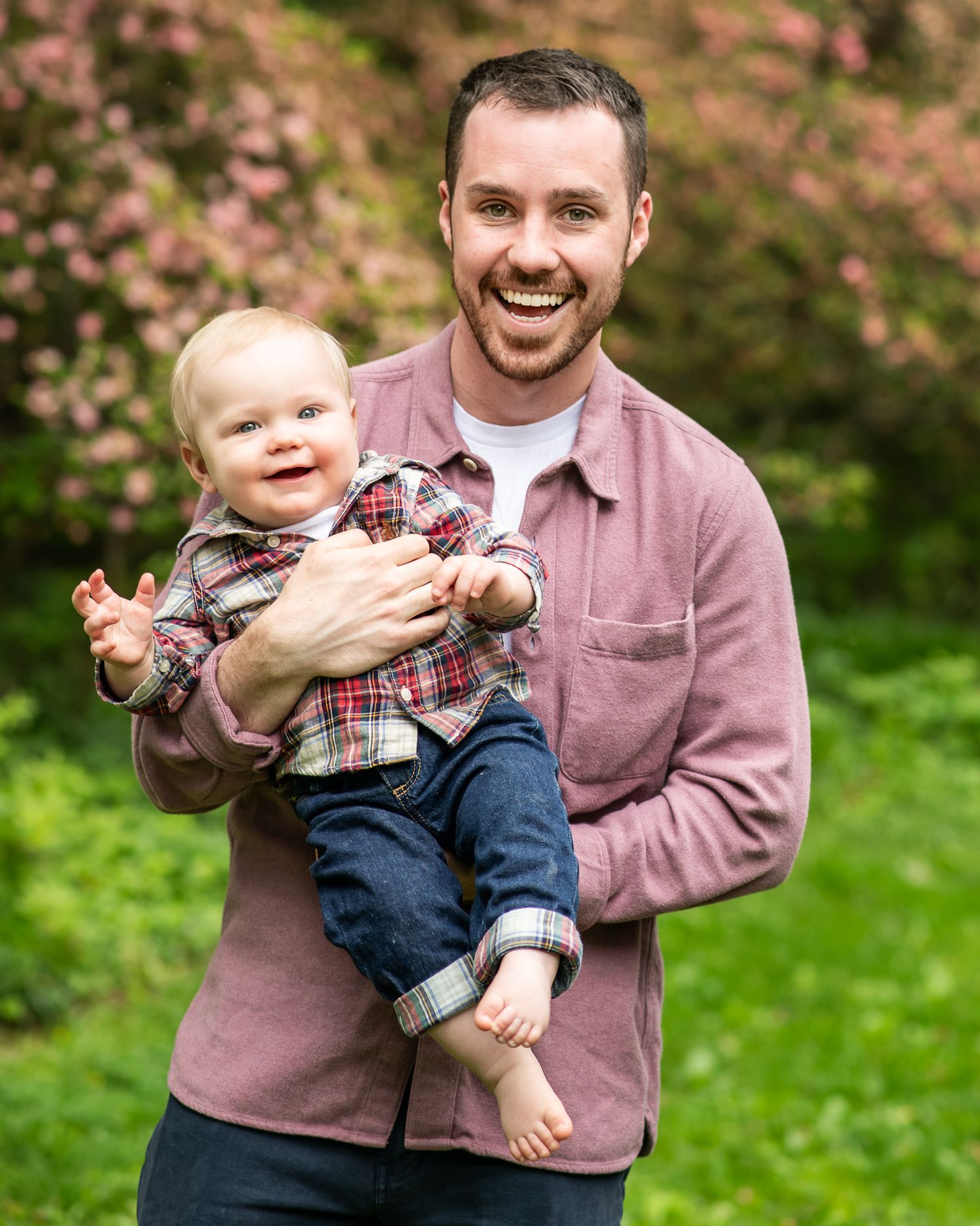 Smiling man holding a happy baby in plaid shirt outdoors with pink flowering trees in background.