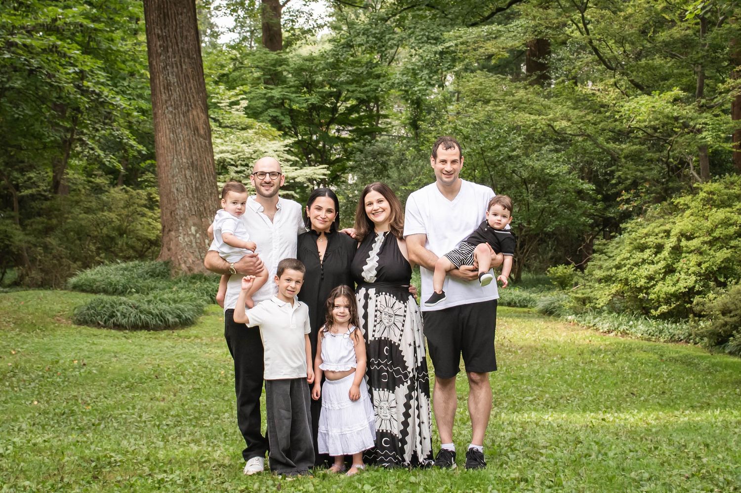A smiling multigenerational family poses together outdoors on a lush green lawn surrounded by trees.