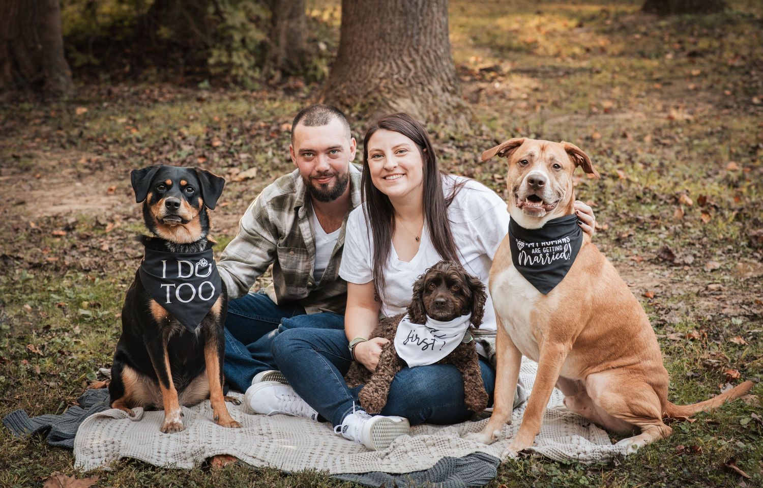 Family sits on blanket with their three dogs wearing matching bandanas in autumn setting.