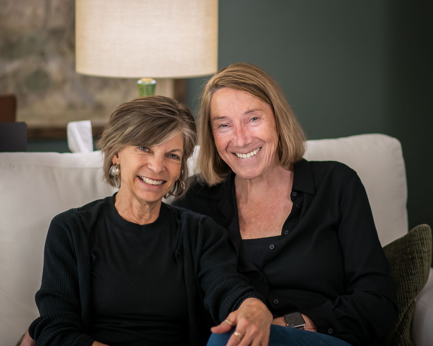 Two smiling women in black tops sitting together on a white couch in a warmly lit room.