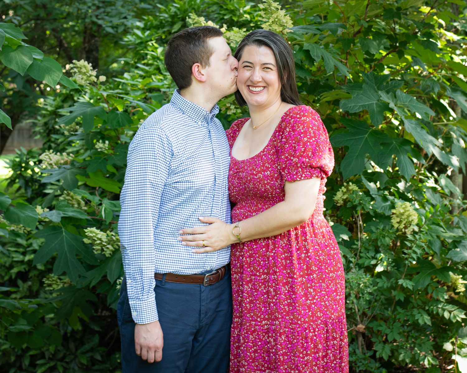 A couple smiles together outdoors, man kissing woman's cheek, woman in pink floral dress, surrounded by lush green foliage.