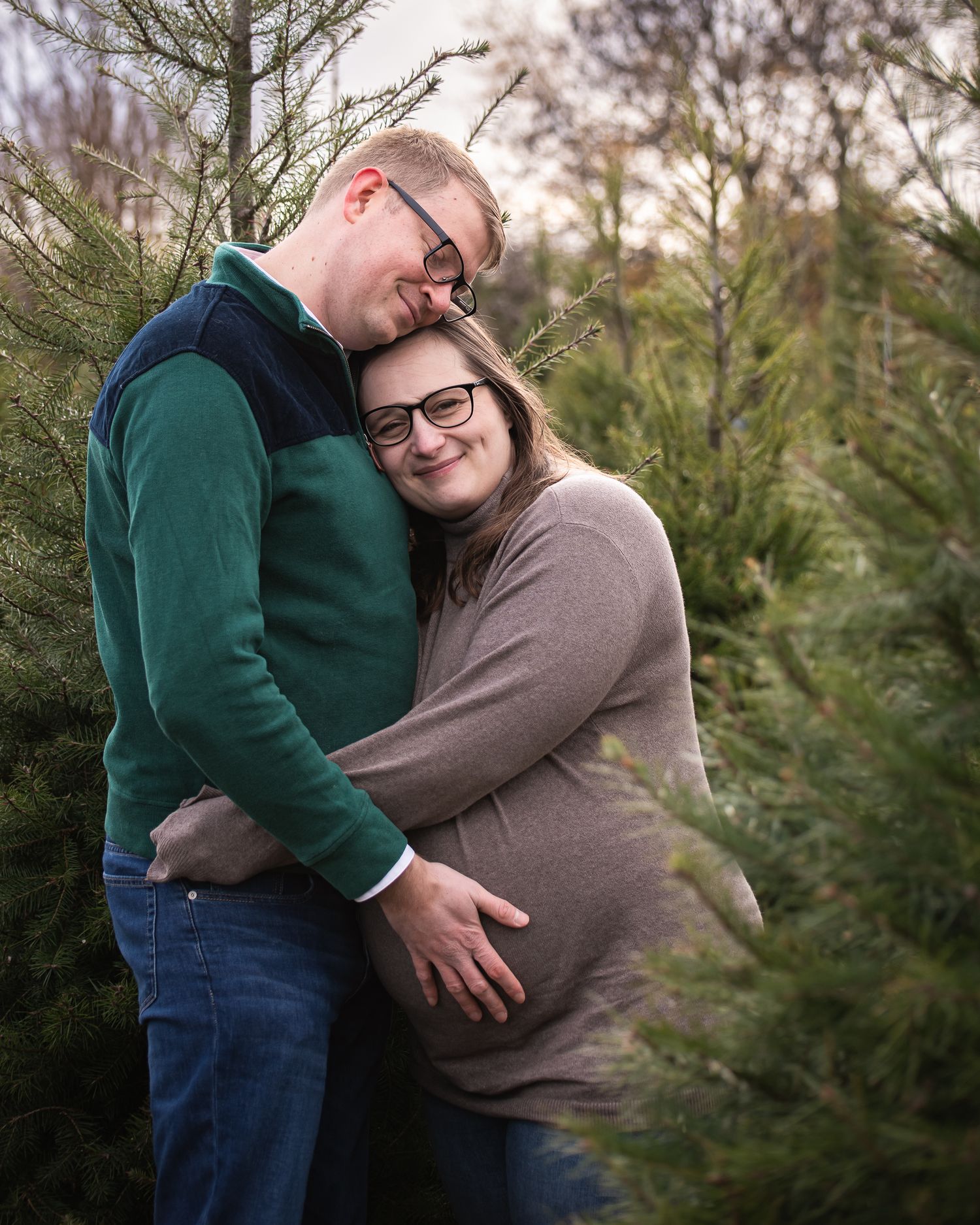 Couple embraces lovingly among evergreen trees at a Christmas tree farm during a maternity photo session.