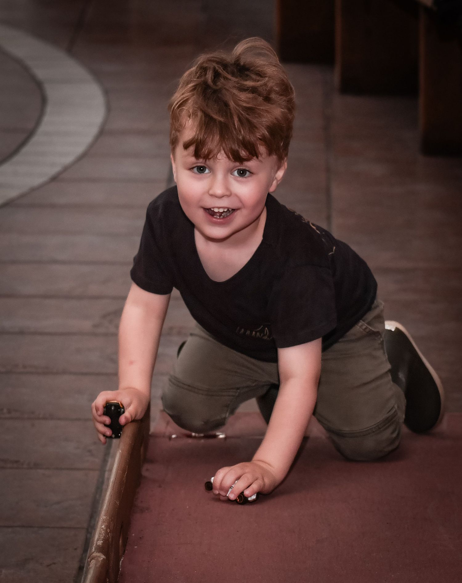A child crawling on the floor with a playful expression wearing a black t-shirt and khaki pants.