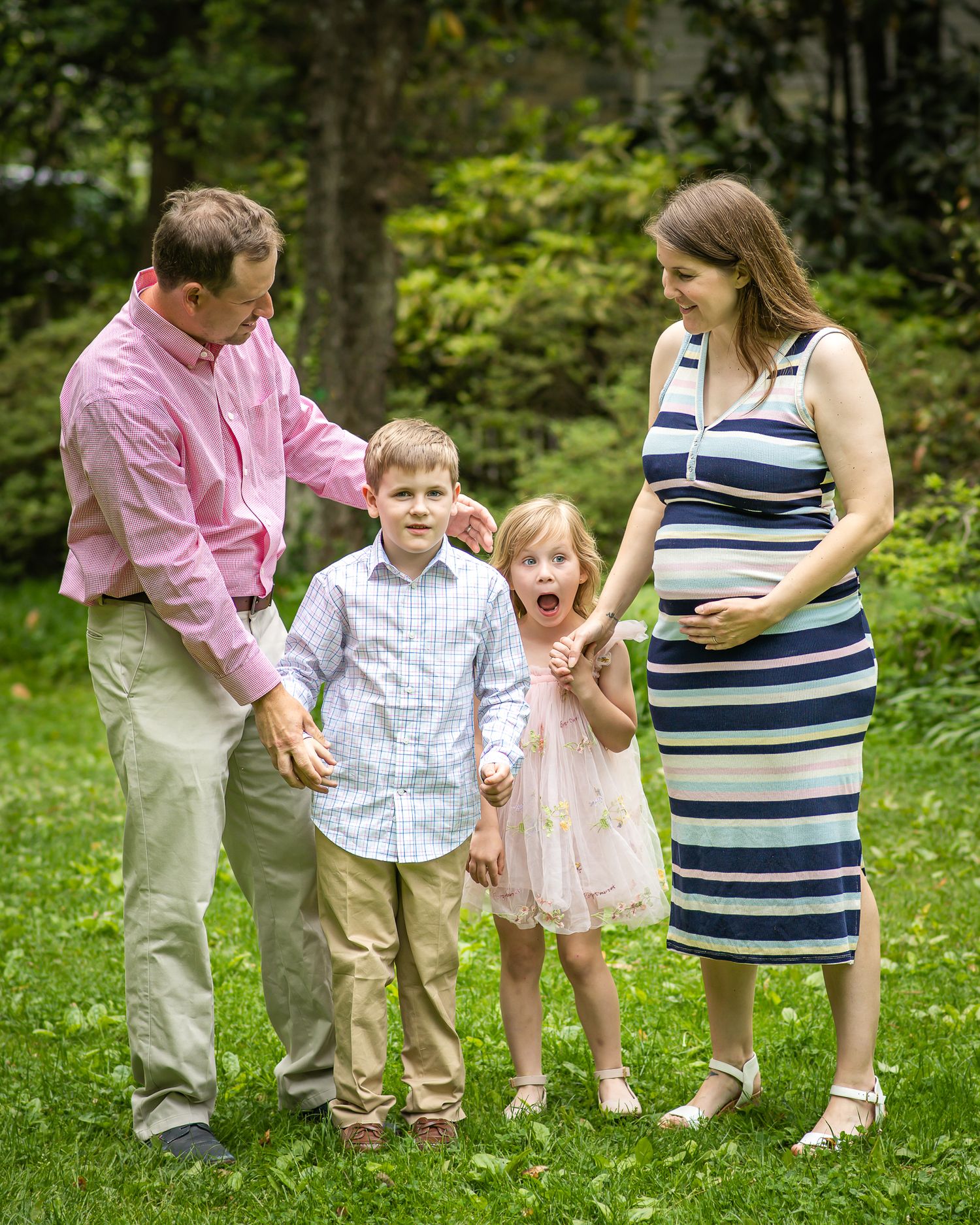 Pregnant woman and husband pose outdoors with young boy and girl on green lawn for family portrait.