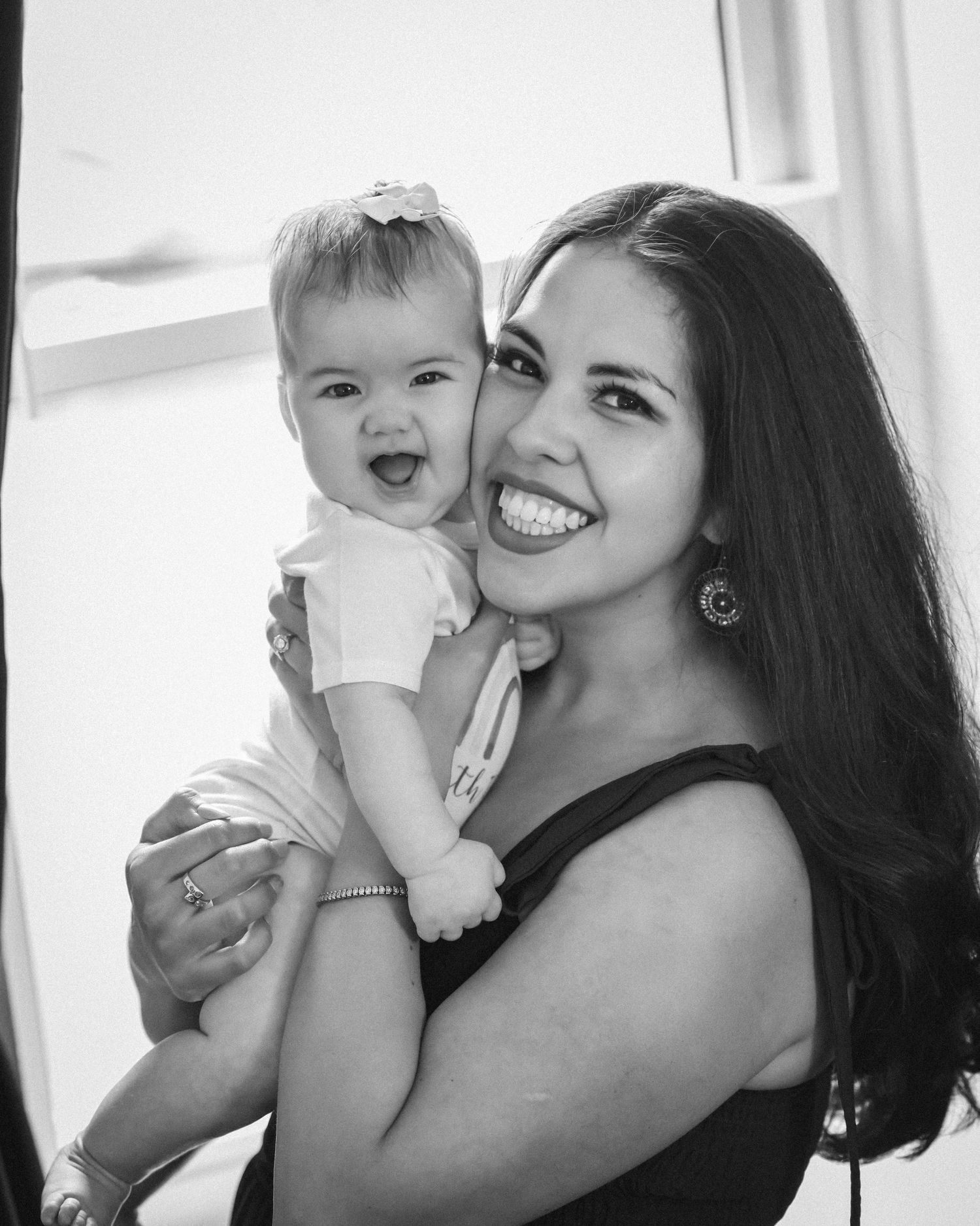 Black and white photo of a smiling woman holding a happy baby girl with a bow in her hair.