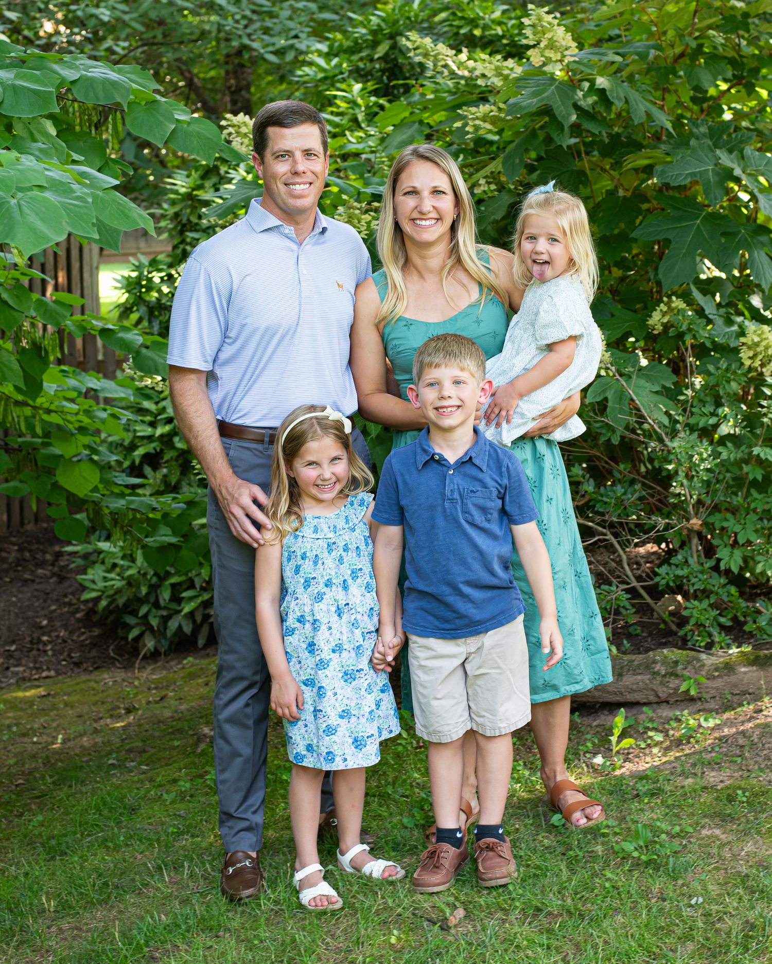 Happy family of five posing outdoors in a lush green garden, parents with three young children smiling.