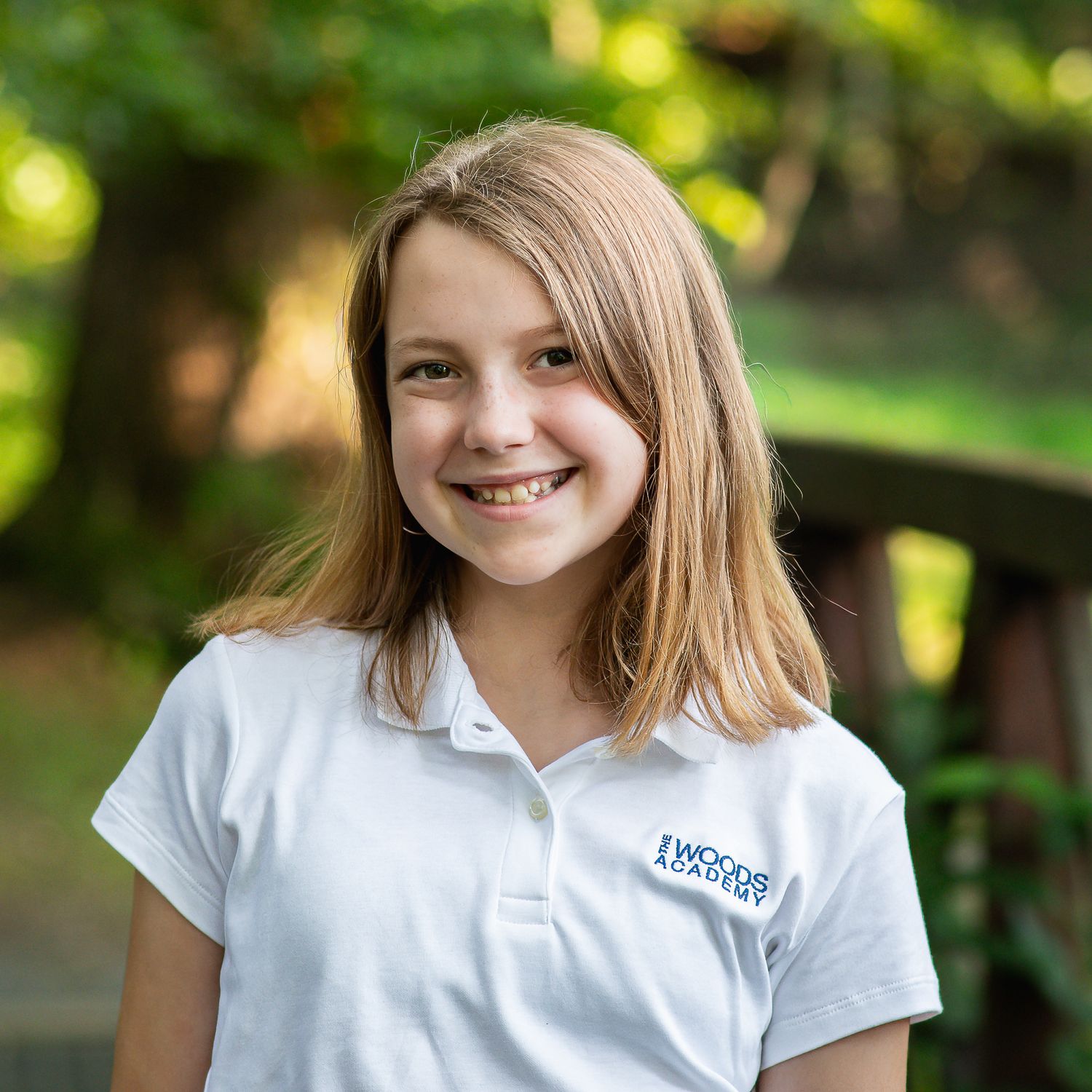 Smiling young girl with blonde hair wearing a white polo shirt outdoors in a lush green setting.