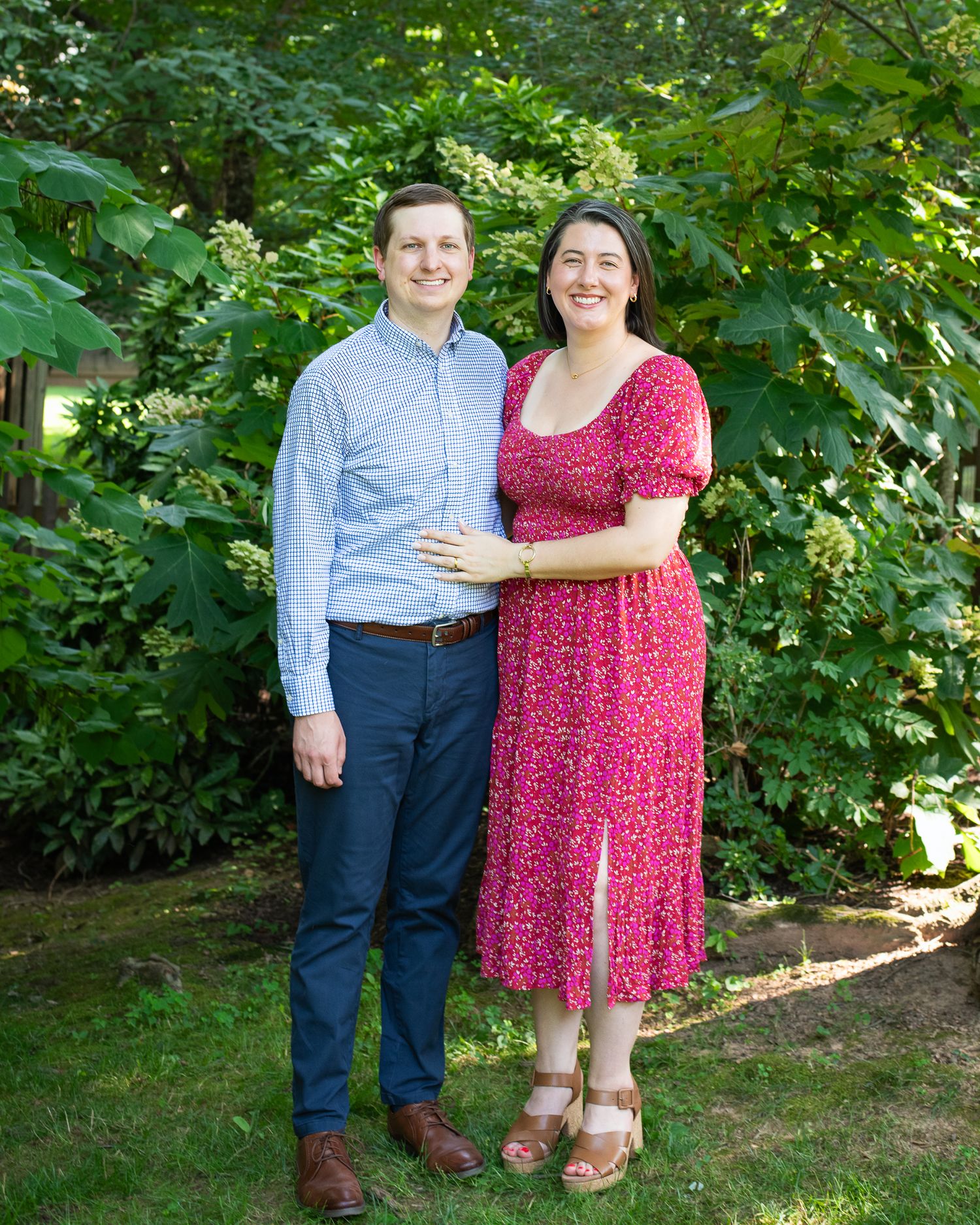 A smiling couple poses together in a lush green garden, man in blue shirt and woman in red floral dress.