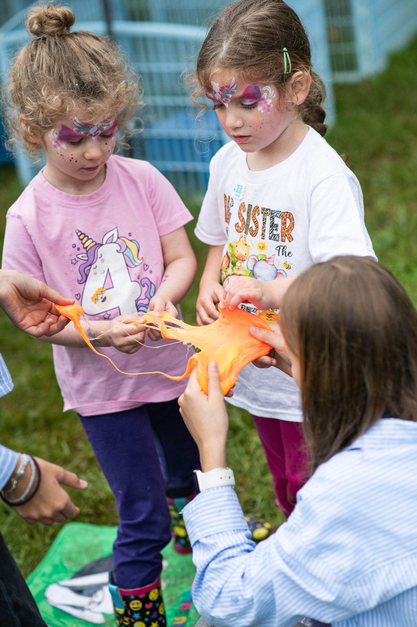 Two girls with face paint play with orange slime at an outdoor birthday party activity.
