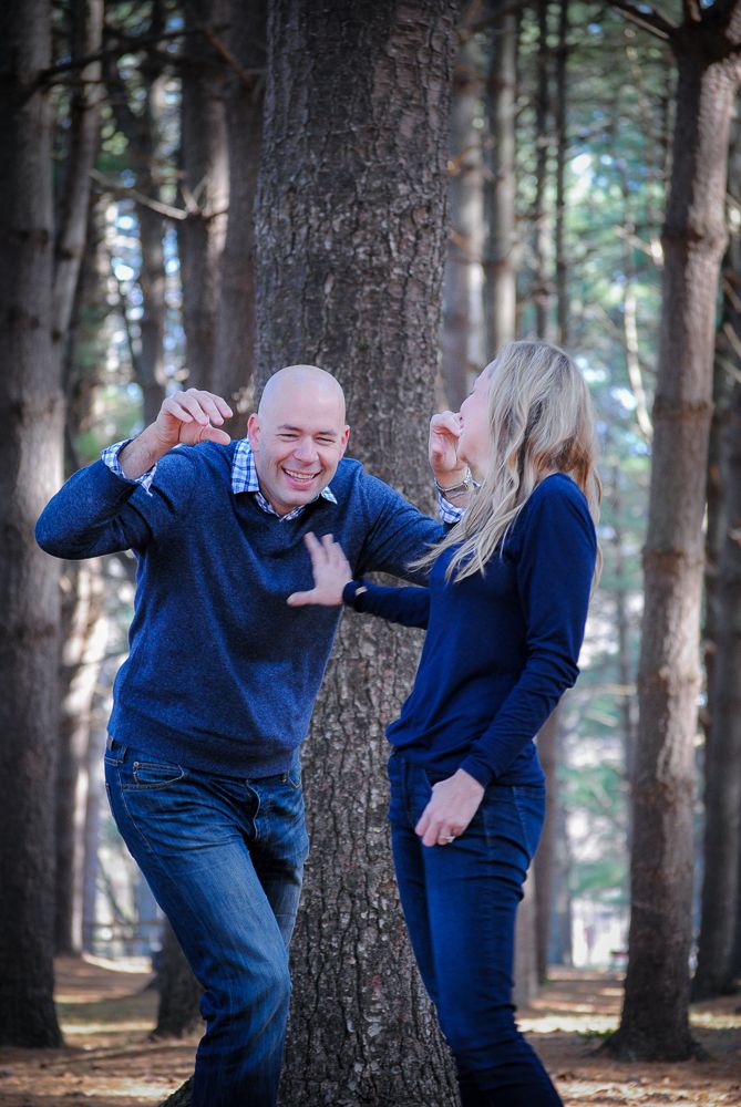 A laughing bald man and blonde woman in blue sweaters playfully pose among tall pine trees outdoors.