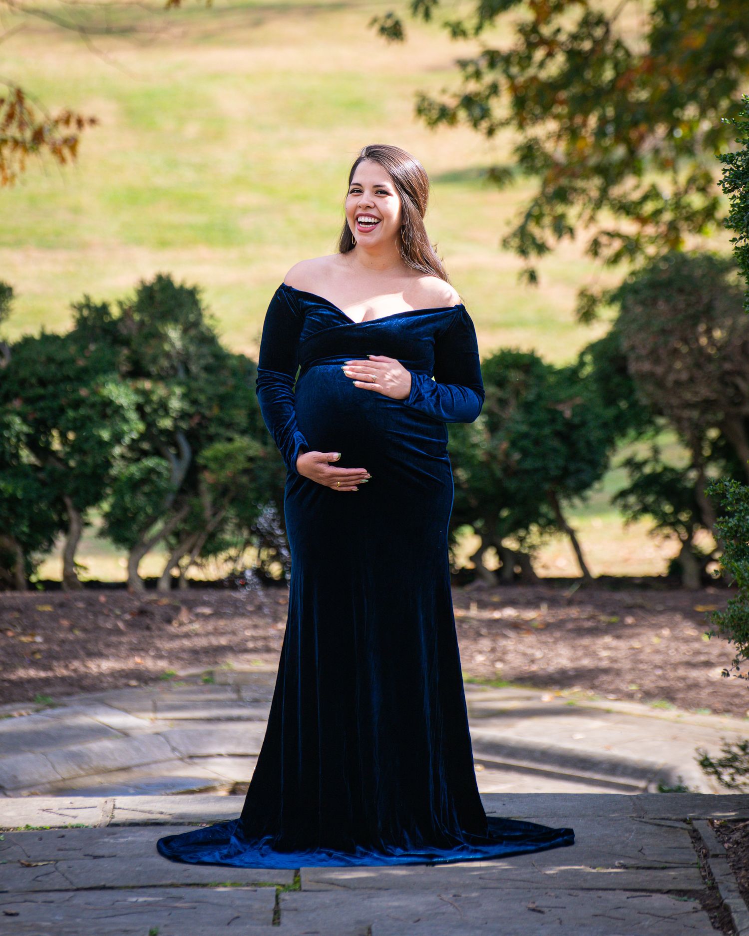 Smiling pregnant woman in elegant navy blue velvet off-shoulder maternity gown posing outdoors in a lush garden.
