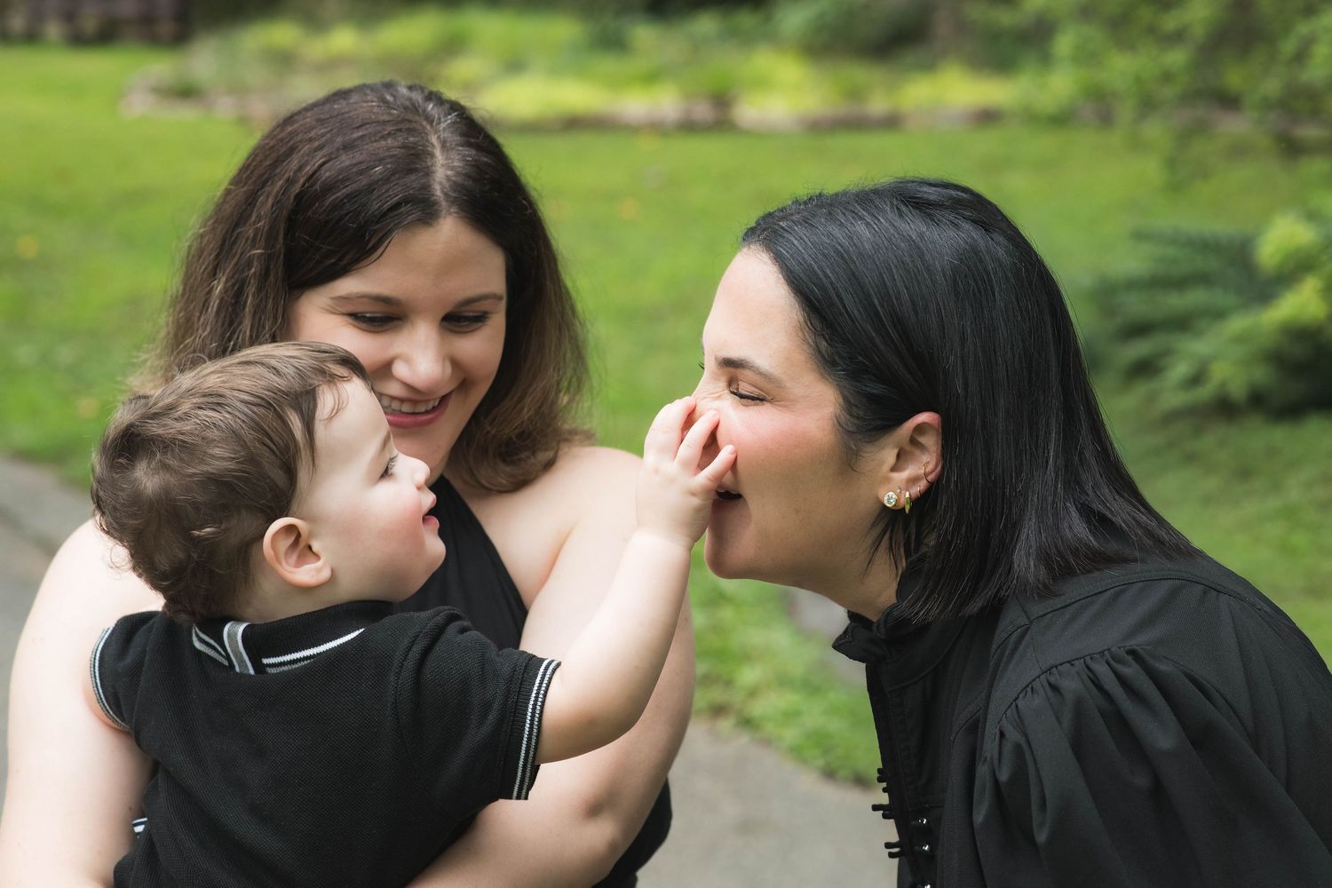 Two women and a baby share a joyful moment outdoors, with one woman kissing the infant's hand.