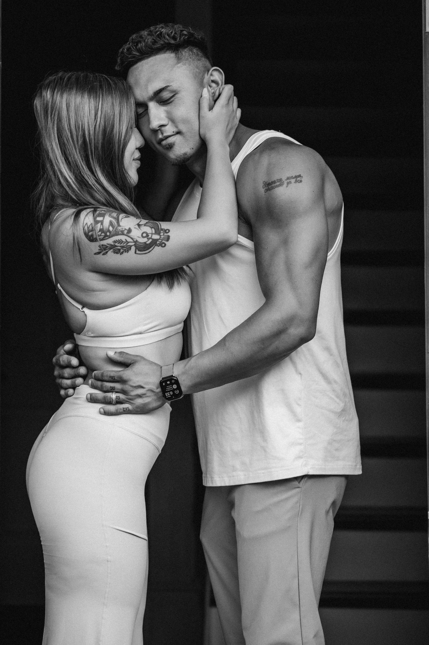 Black and white photo of a tattooed couple embracing intimately, the woman in a white dress, the man in a sleeveless shirt.