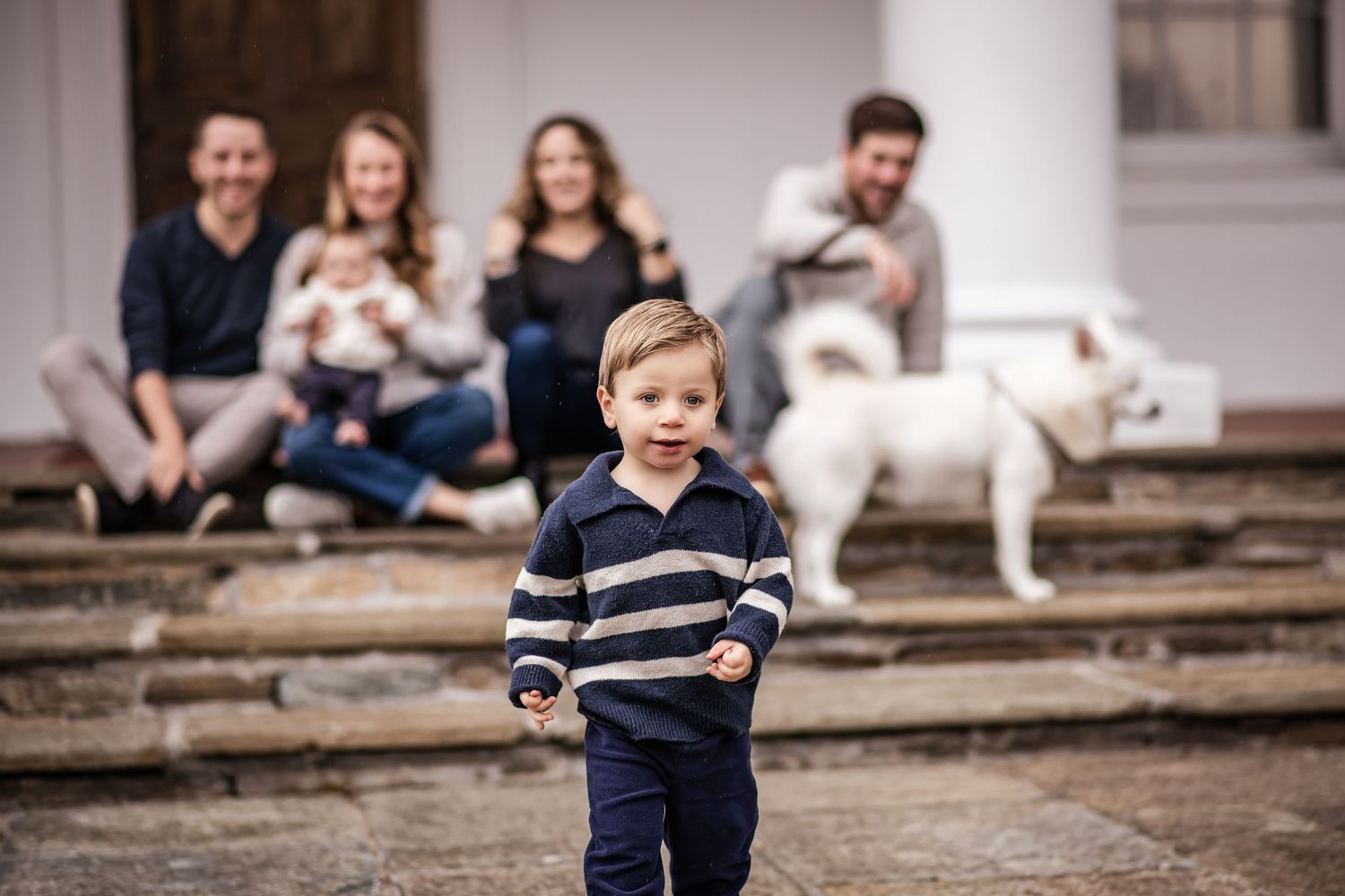 A young child in a navy striped shirt plays outdoors on a rustic porch with family and a white dog visible in background.