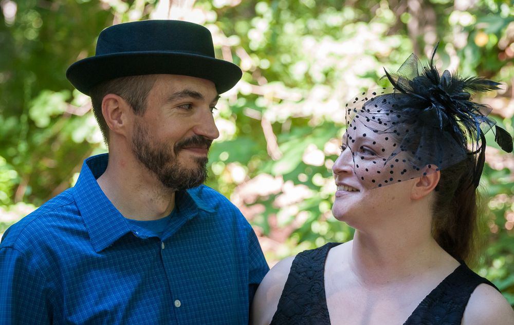 A couple at an outdoor event, with one wearing a black hat and the other in a decorative face mask.