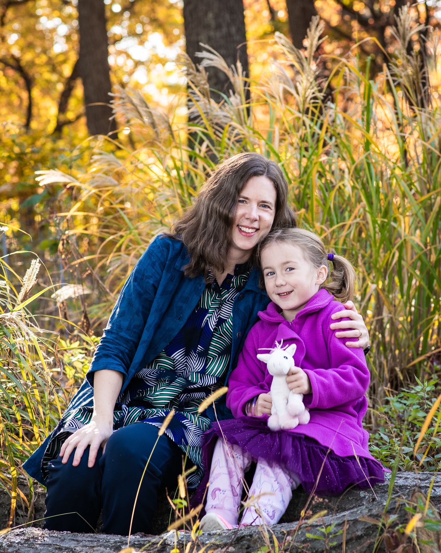 A smiling woman and young girl holding a stuffed unicorn pose together outdoors among tall golden grasses in autumn.