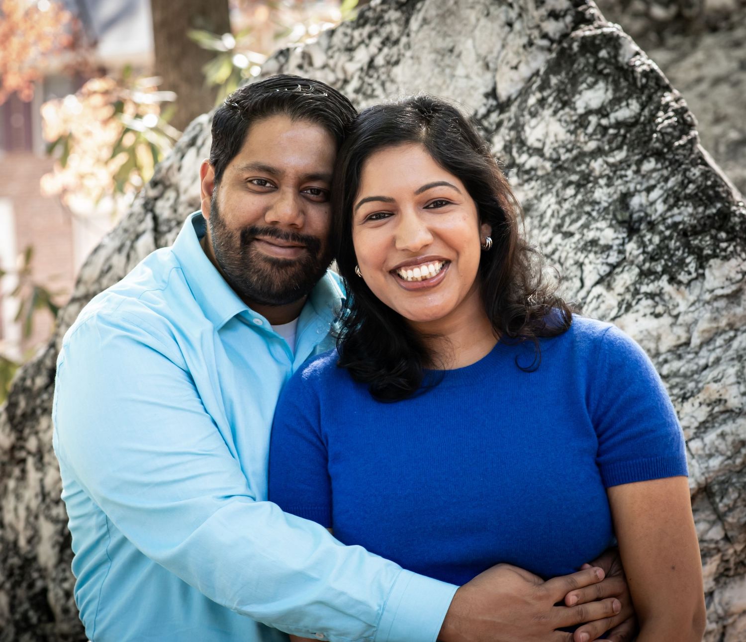 A couple in blue outfits share loving embraces while posing for outdoor portrait photos against a natural background.