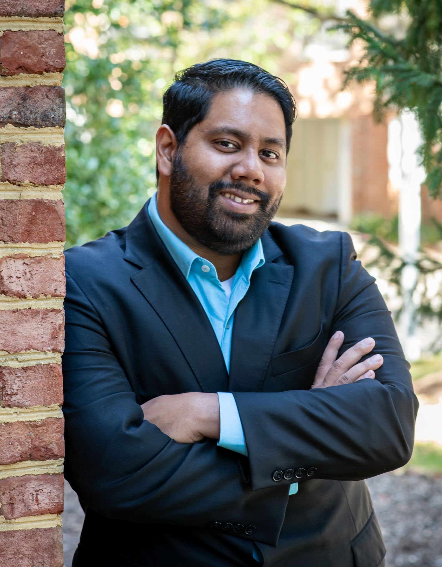Professional wearing a dark suit and light blue shirt poses confidently against a brick wall in an outdoor setting.