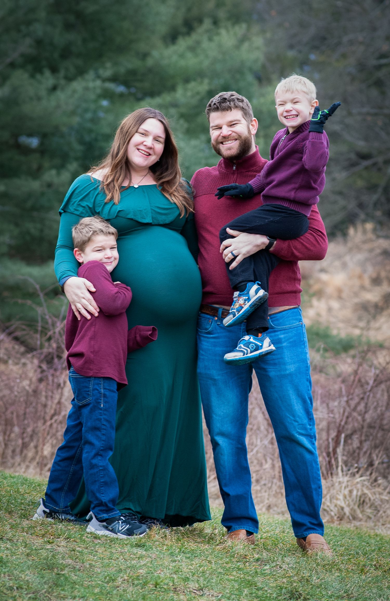 A small family poses together outdoors, wearing coordinating burgundy and teal outfits for a maternity photo session.