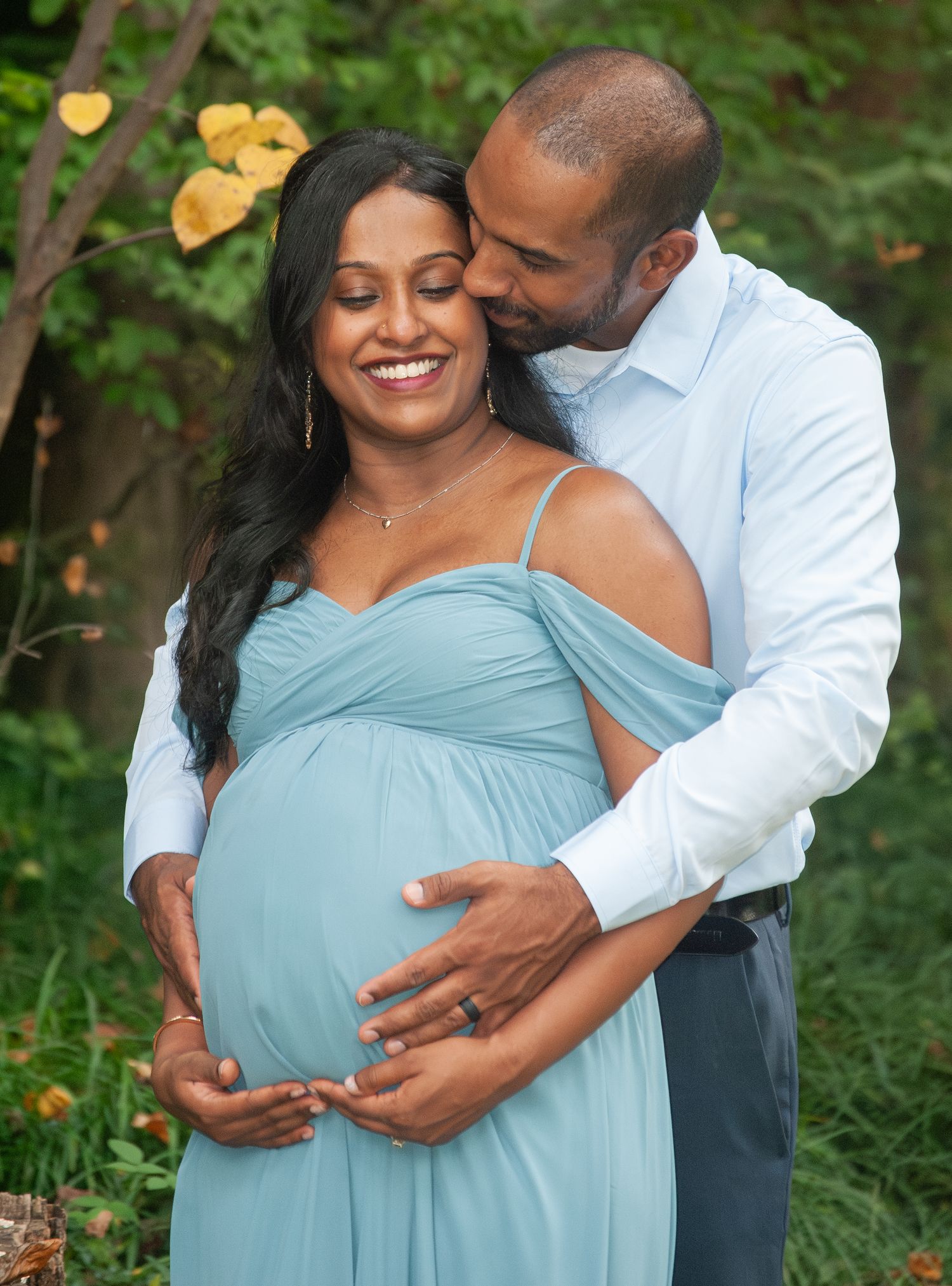 Pregnant woman in blue gown embraced by partner outdoors during maternity photo session.