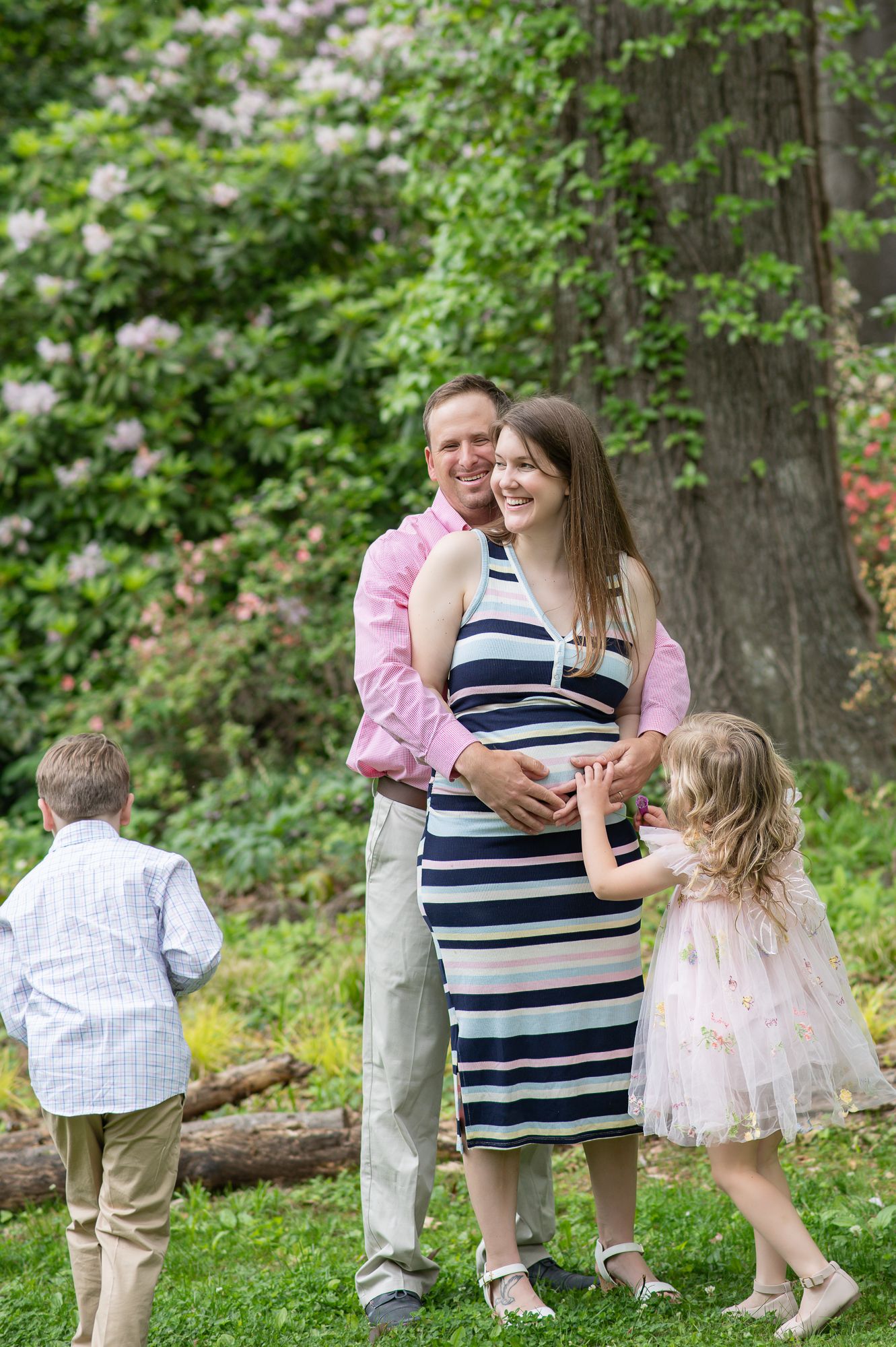 Pregnant woman in striped dress with husband and two children posing outdoors in lush green garden with blooming flowers.