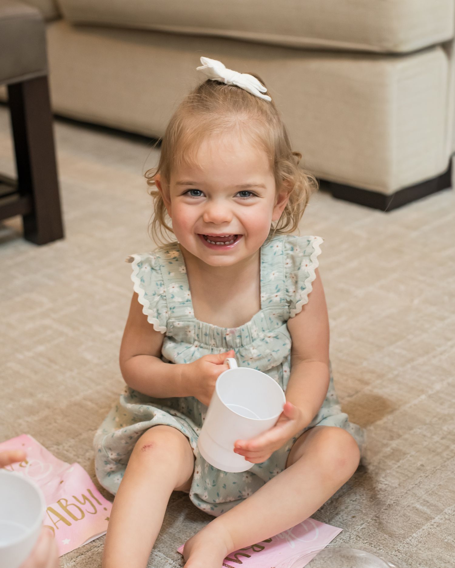 Smiling toddler girl in floral dress with white bow holding a cup, sitting on carpet.