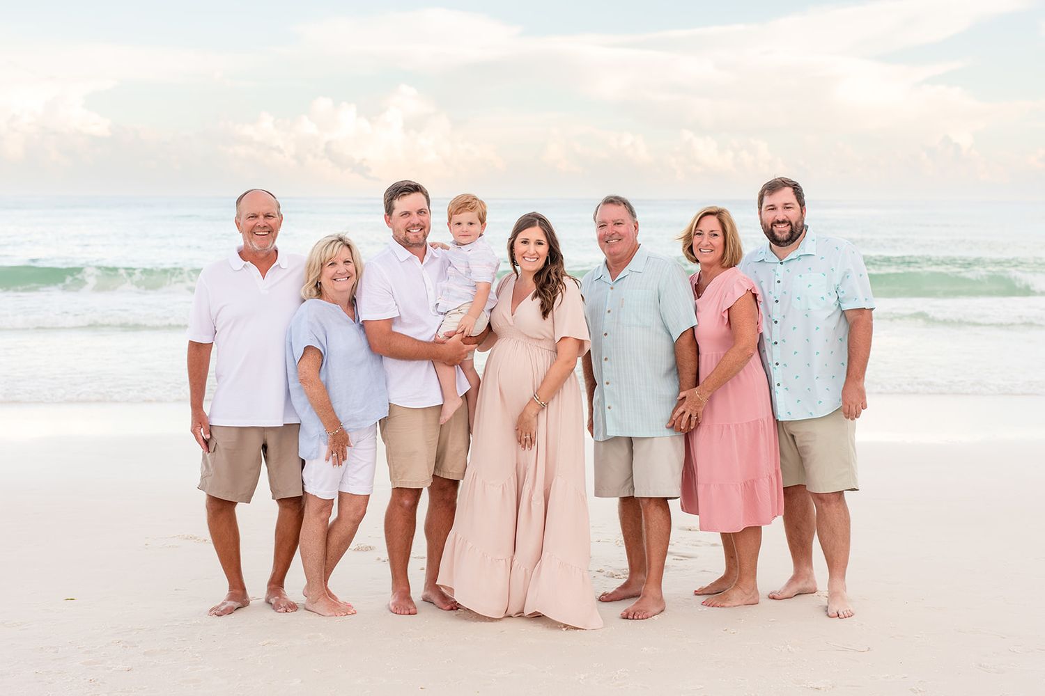 A group in white and neutral colored clothing poses together on a sunny beach at sunset.