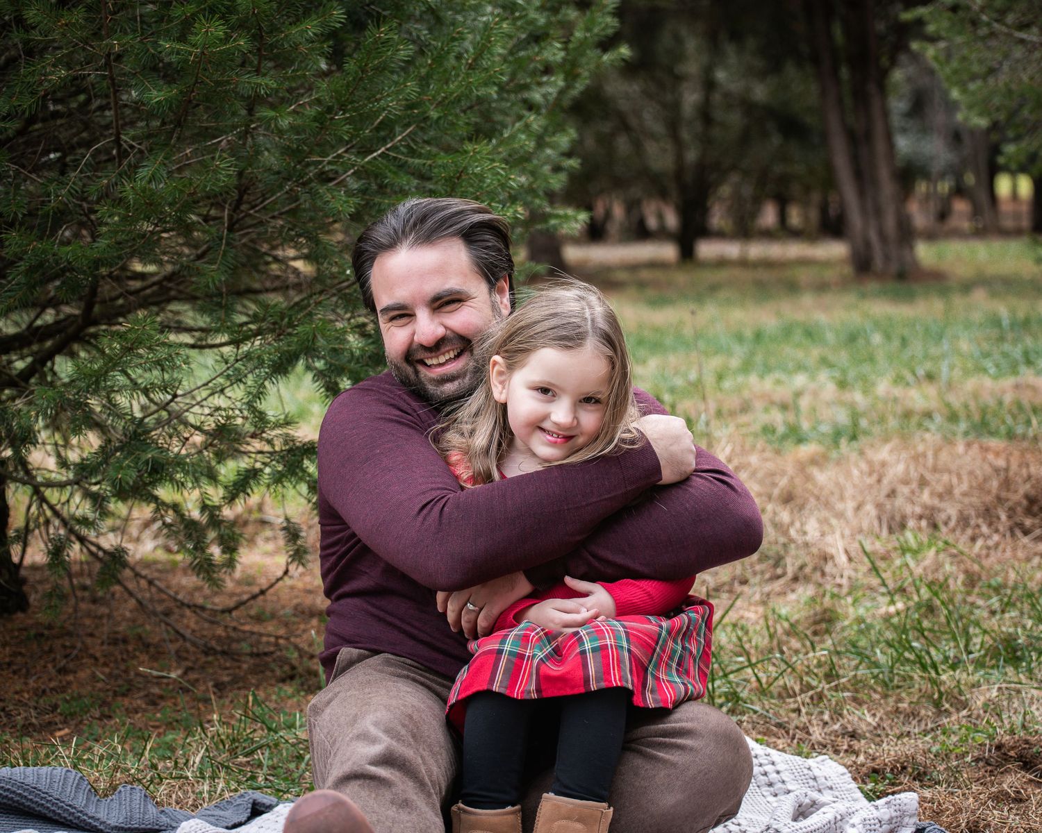 A couple in burgundy sweaters sits together on a blanket during an outdoor fall photoshoot in a wooded area.