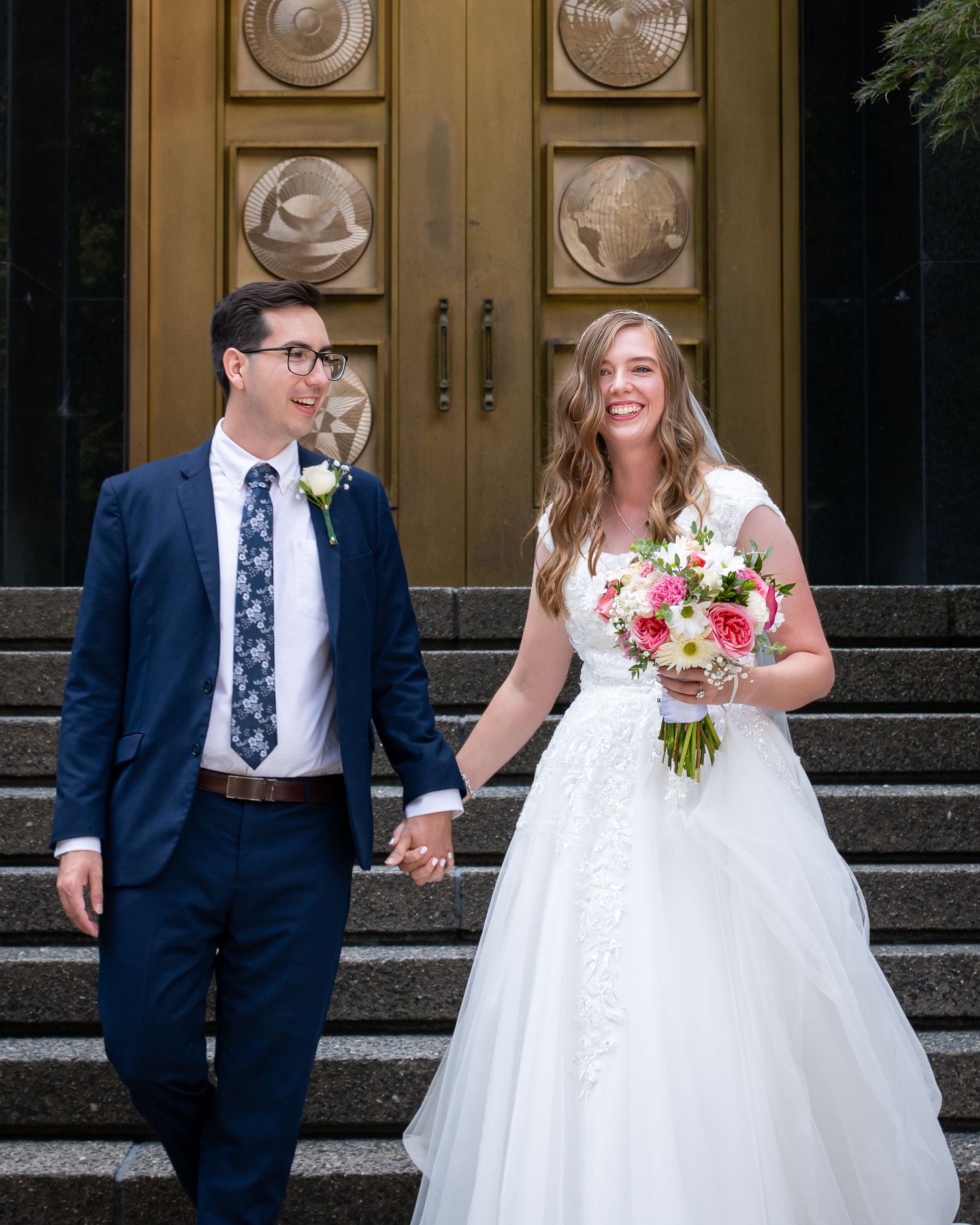 Smiling newlyweds hold hands on church steps, bride in white gown with bouquet, groom in navy suit with floral tie.