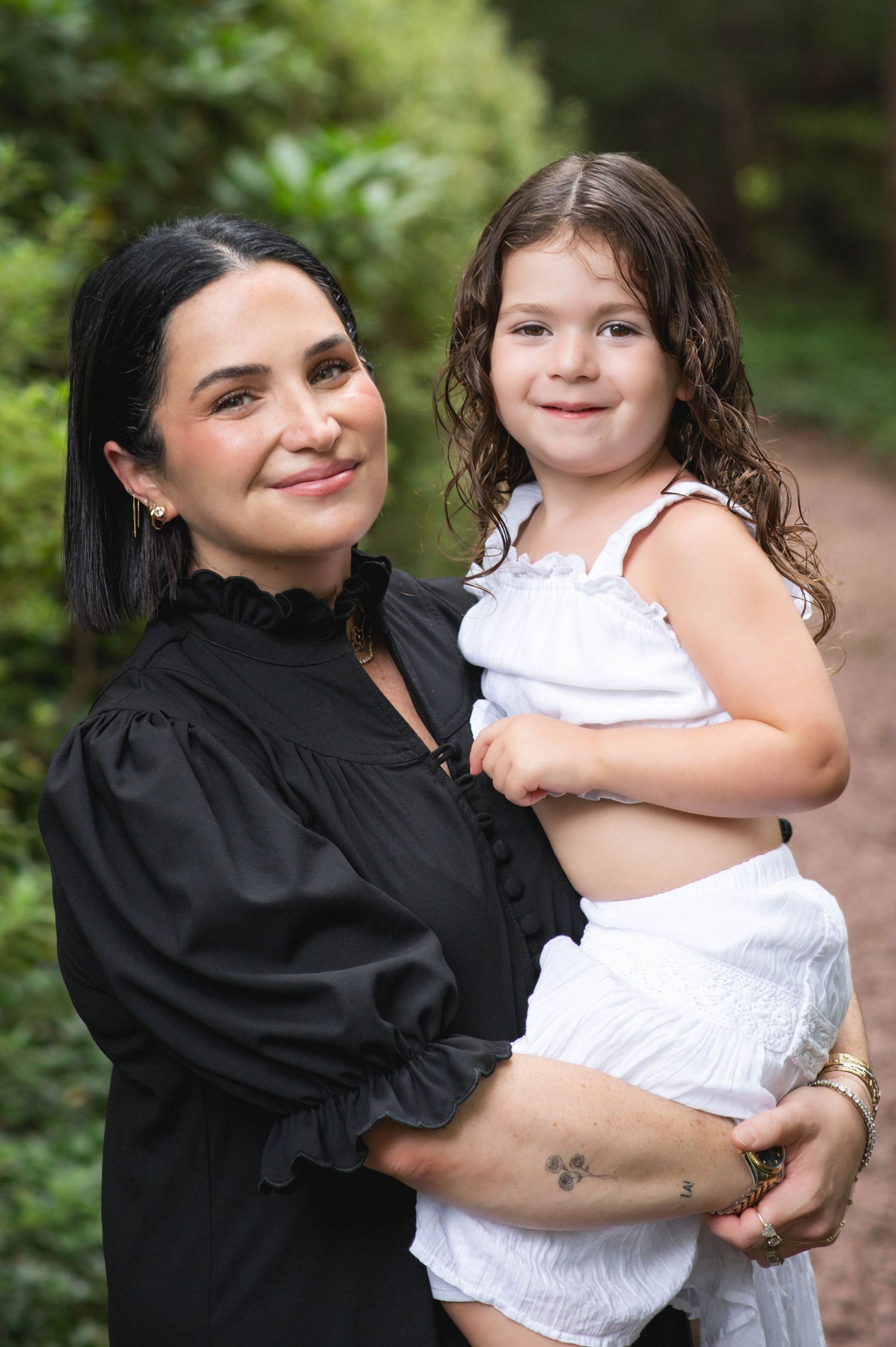 A woman in black dress holds a young girl in white outfit, smiling outdoors among green trees.