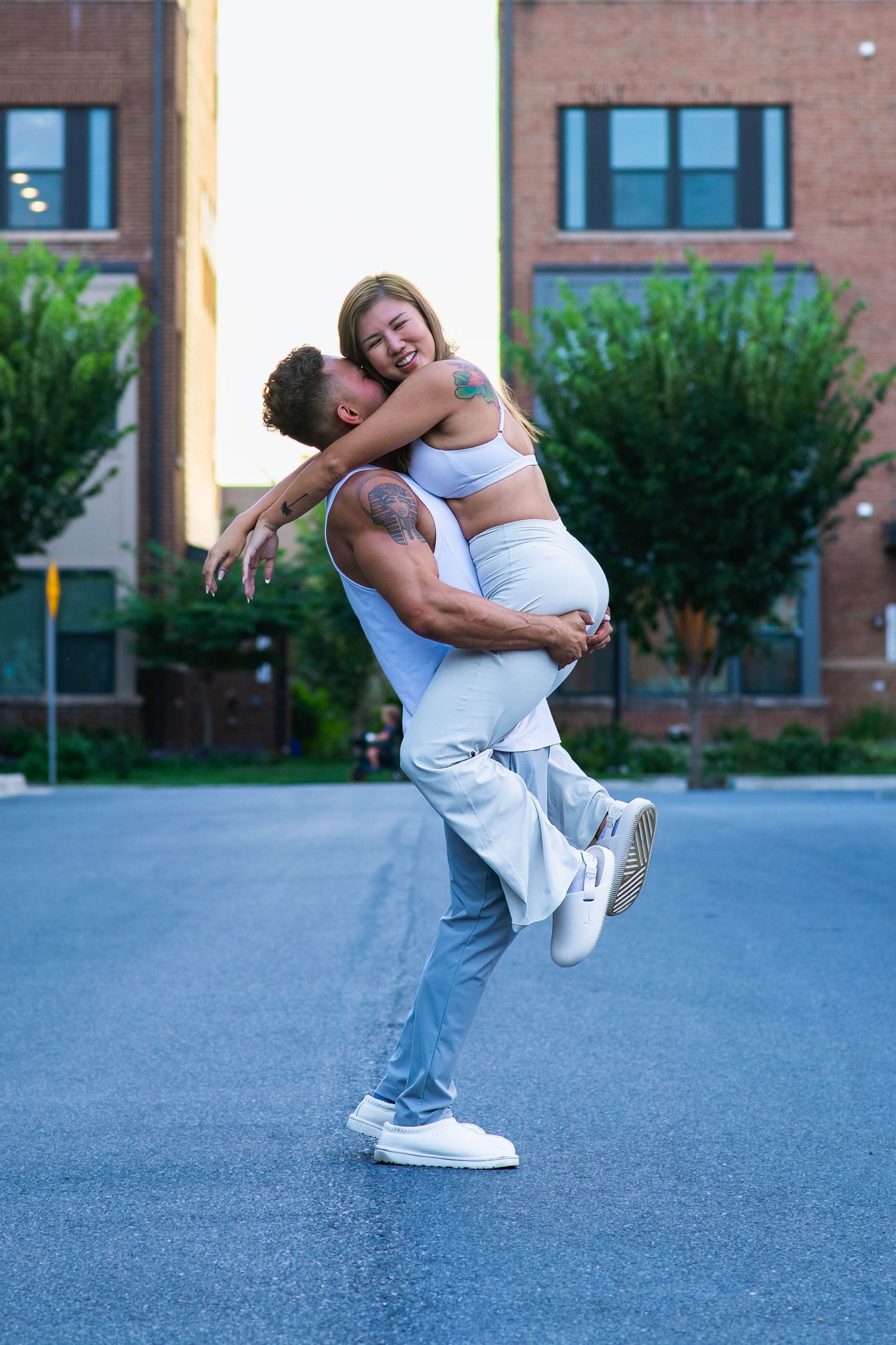A couple dances joyfully outdoors, man lifting woman in matching light blue outfits near brick building.