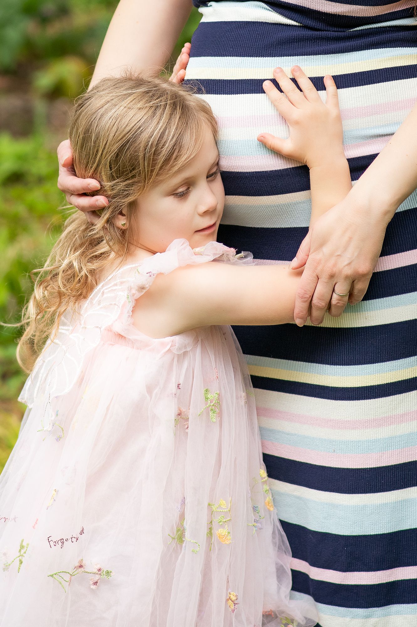 Young girl in pink tulle dress hugging pregnant mother's belly outdoors in garden setting.