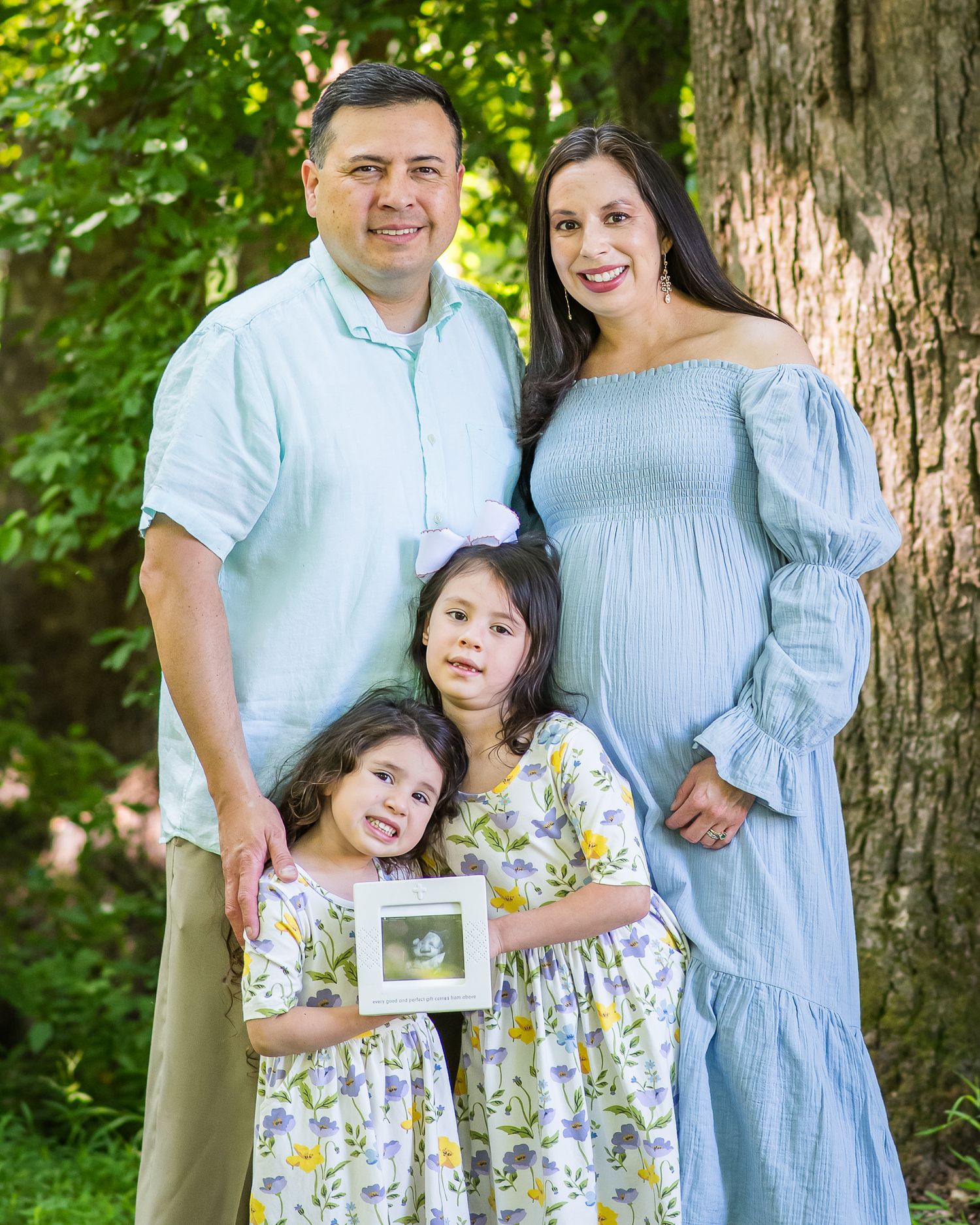 Pregnant woman in blue dress poses with husband and two daughters holding ultrasound photo outdoors.